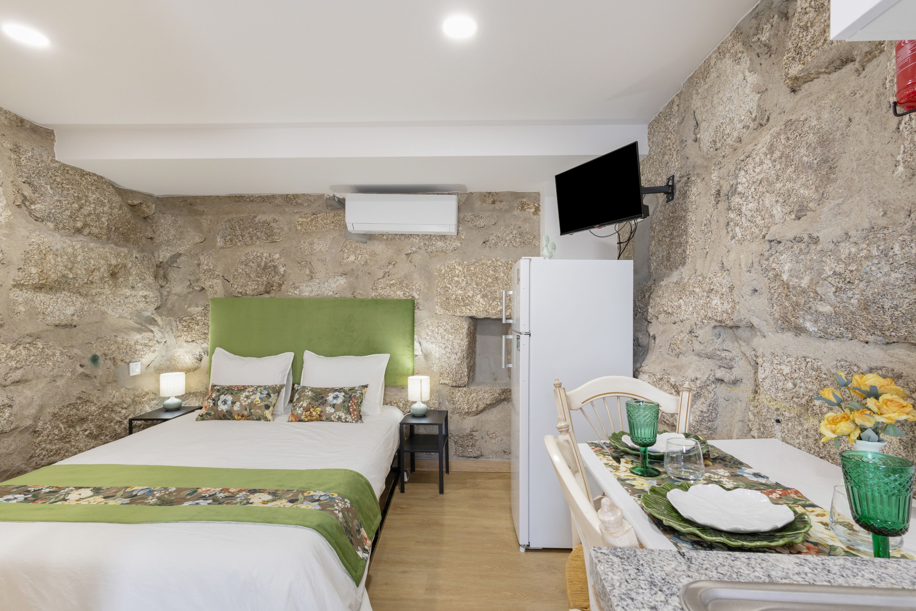 Un dormitorio con cama, mesitas de noche, televisor, refrigerador y un rincón de comedor con mesa puesta para dos.