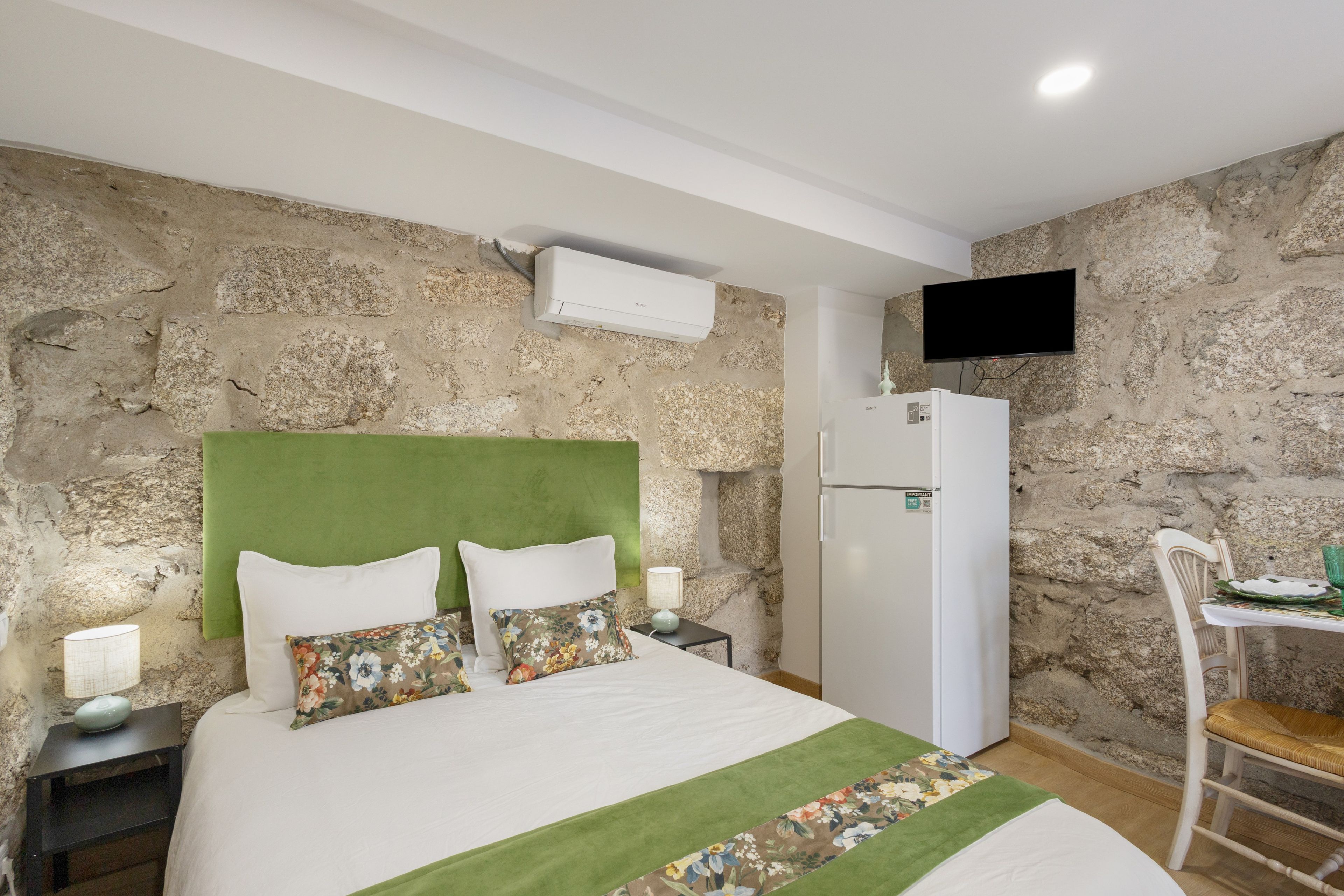 Un dormitorio con pared de piedra, una cama con almohadas florales, un aire acondicionado blanco, un televisor de pantalla plana montado en la pared y una pequeña mesa con una silla.