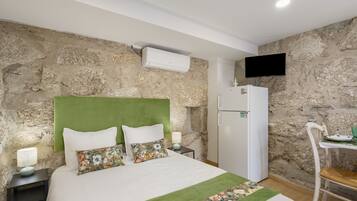 Un dormitorio con pared de piedra, una cama con almohadas florales, un aire acondicionado blanco, un televisor de pantalla plana montado en la pared y una pequeña mesa con una silla.