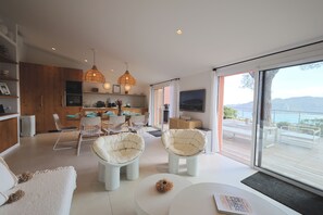 A modern living room with a dining area, large windows, and a view of the sea. - U Mulinu | Villa with Ocean Views | Panorama Santa Giulia (Porto-Vecchio)