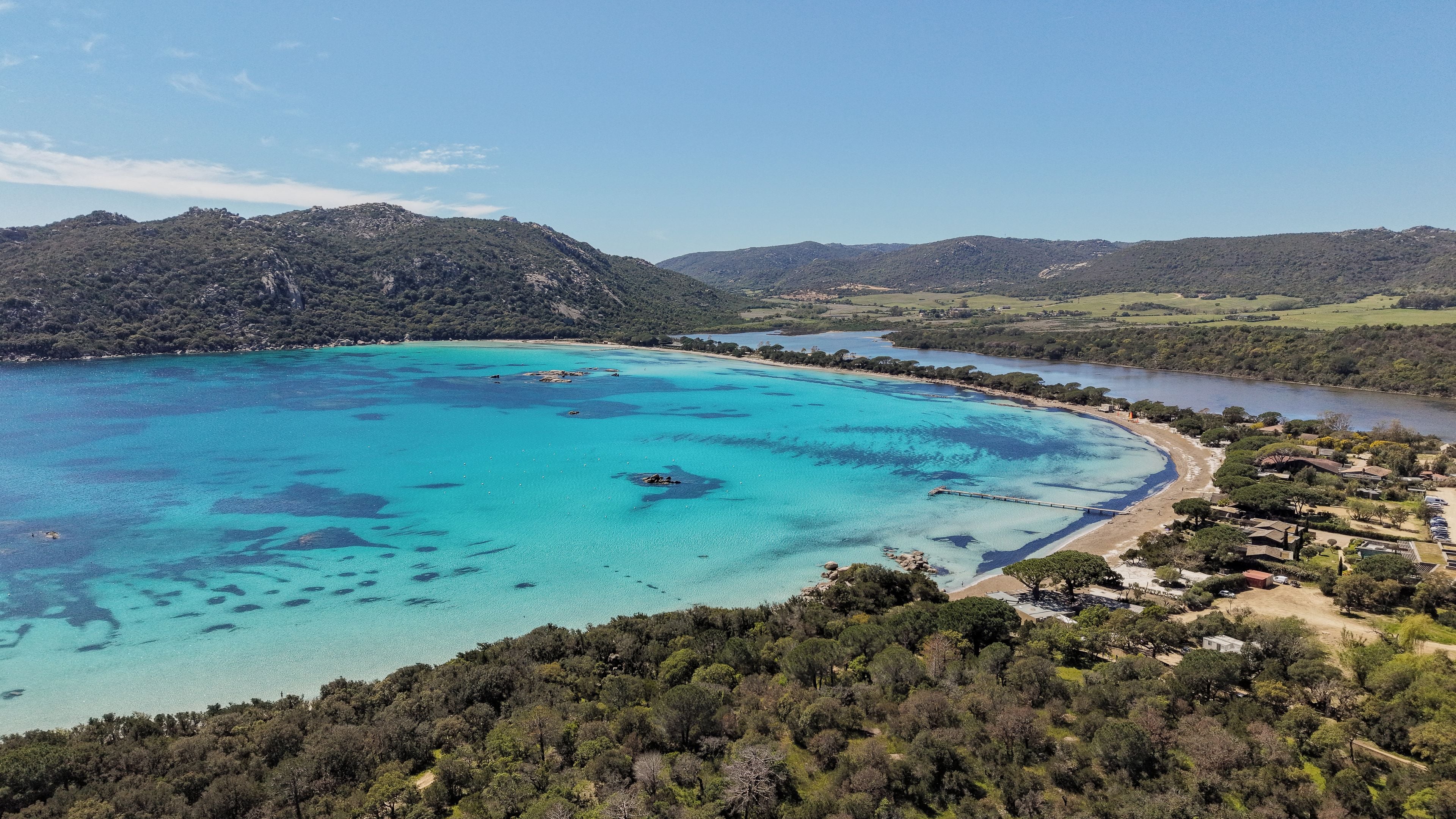 Eine Küstenlandschaft mit klarem türkisblauem Meer, einem Sandstrand und einem bewaldeten Hügel.