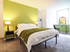 A bed with white linens and a green throw, flanked by two bedside tables with lamps, a desk with a mirror, and a chair. - Flat 1 Greenfields (Barmouth)