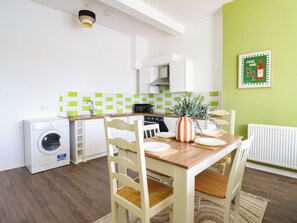 A compact kitchen with a washing machine, a dining table with chairs, and a green wall with a framed picture. - Flat 1 Greenfields (Barmouth)