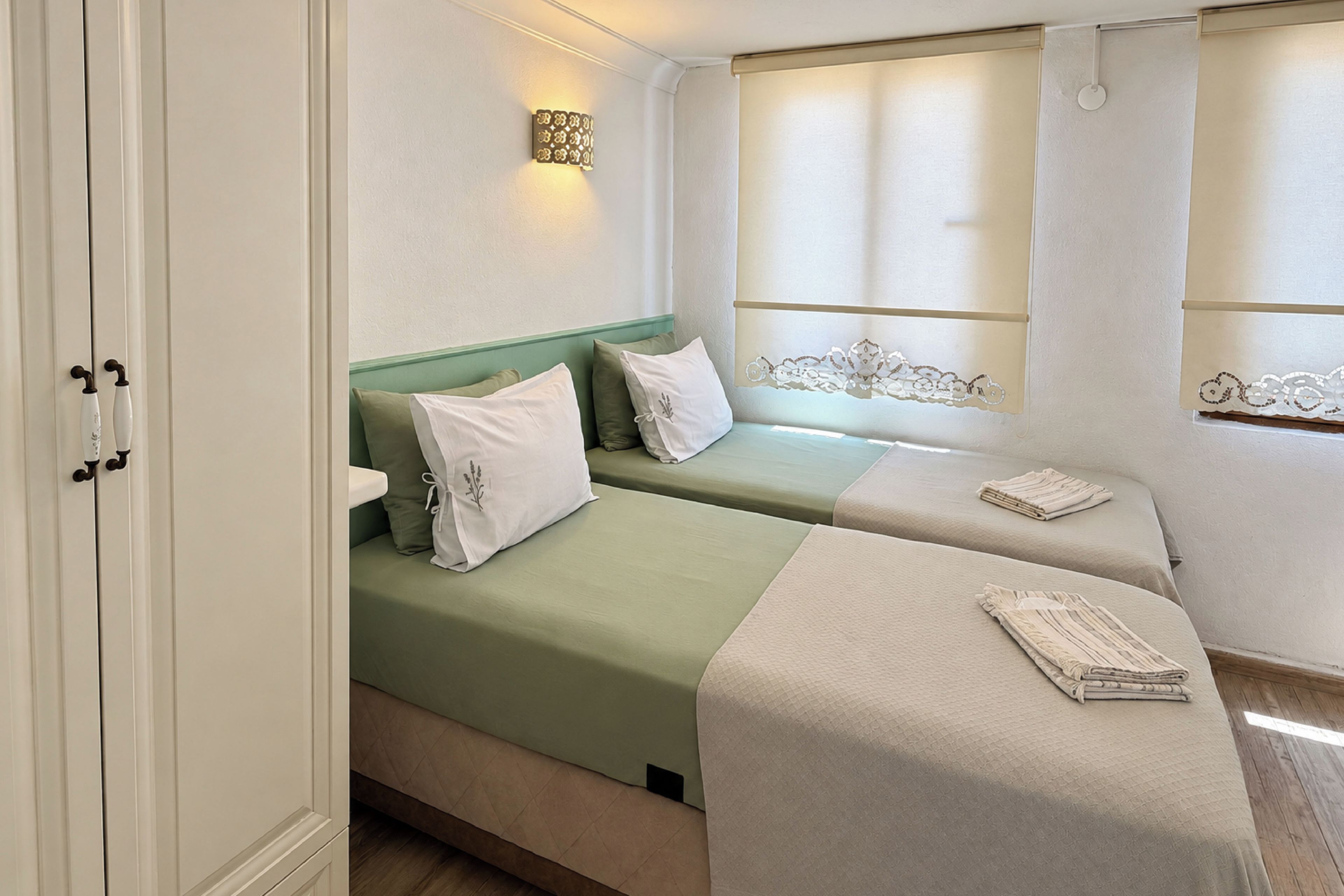 A hotel room with two beds, a window with curtains, a wall-mounted light fixture, and a wooden floor.