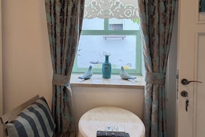 A room with a window, curtains, a sofa, a small stool, and a vase with decorative items on the windowsill. - Villa Teos Sığacık (Seferihisar)