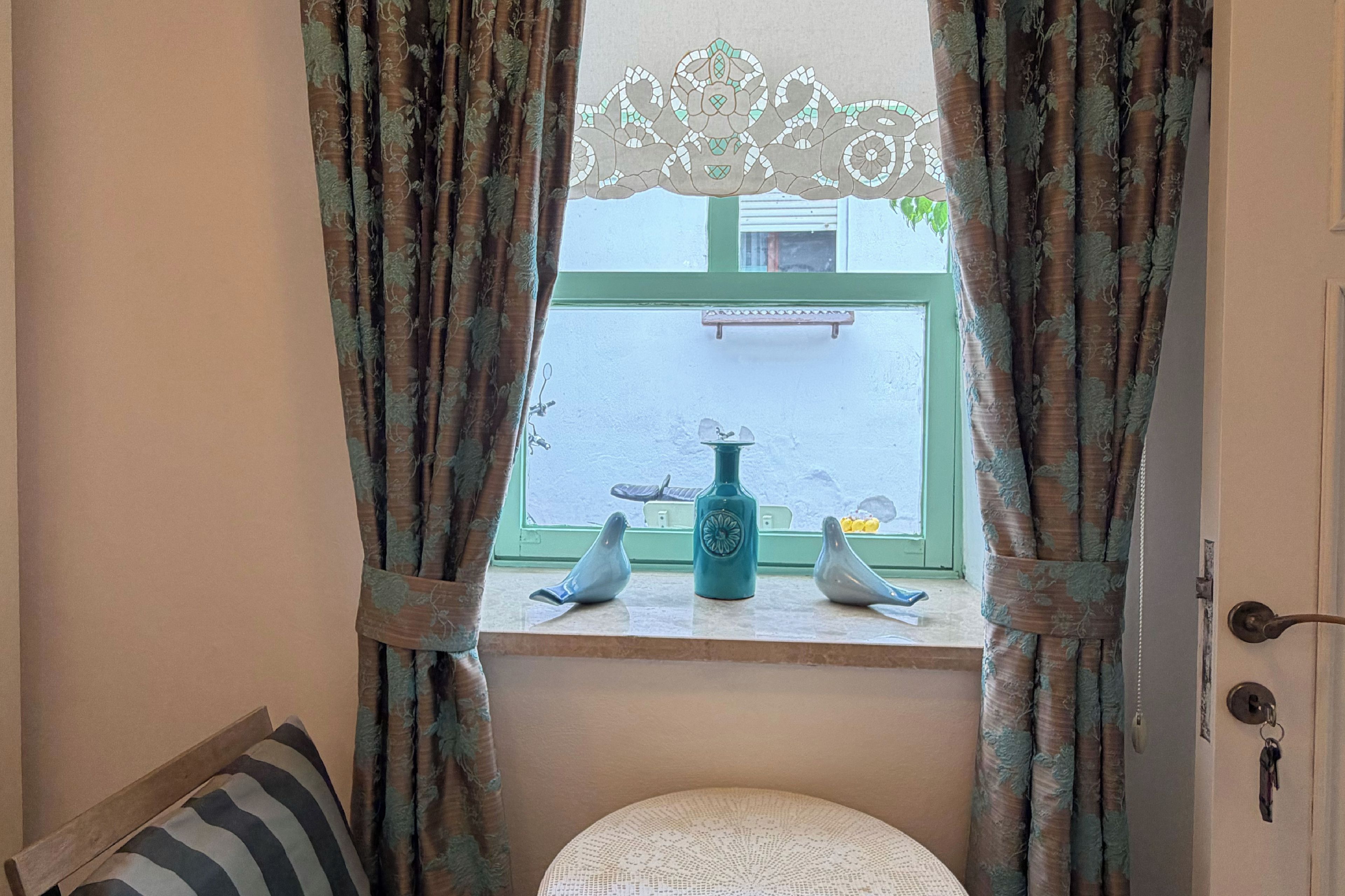 Una habitación con ventana, cortinas, una mesita con adornos y una puerta.