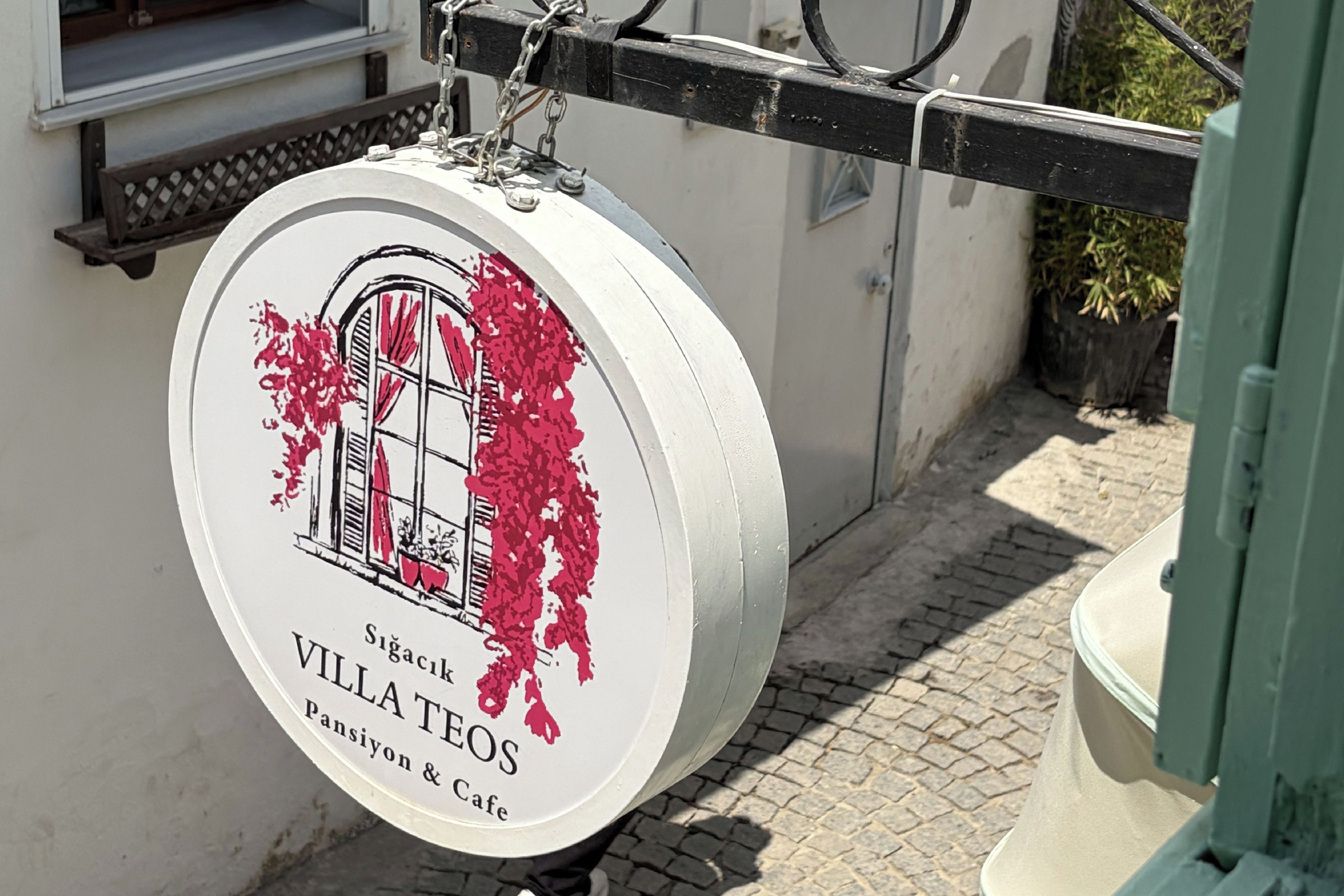 Un letrero redondo con una ilustración de un edificio y el texto 'Villatos Pansiyon & Cafe'.