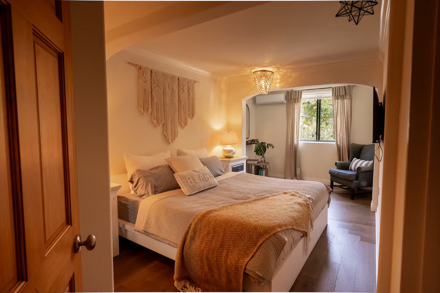Un dormitorio con cama, mesitas de noche, lámpara, una silla y una ventana con cortinas.