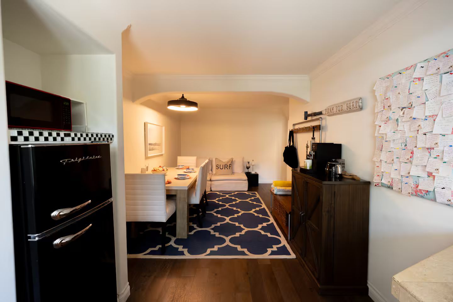 A compact kitchen with a refrigerator, a dining table with chairs, a sofa, and a wall with notes.