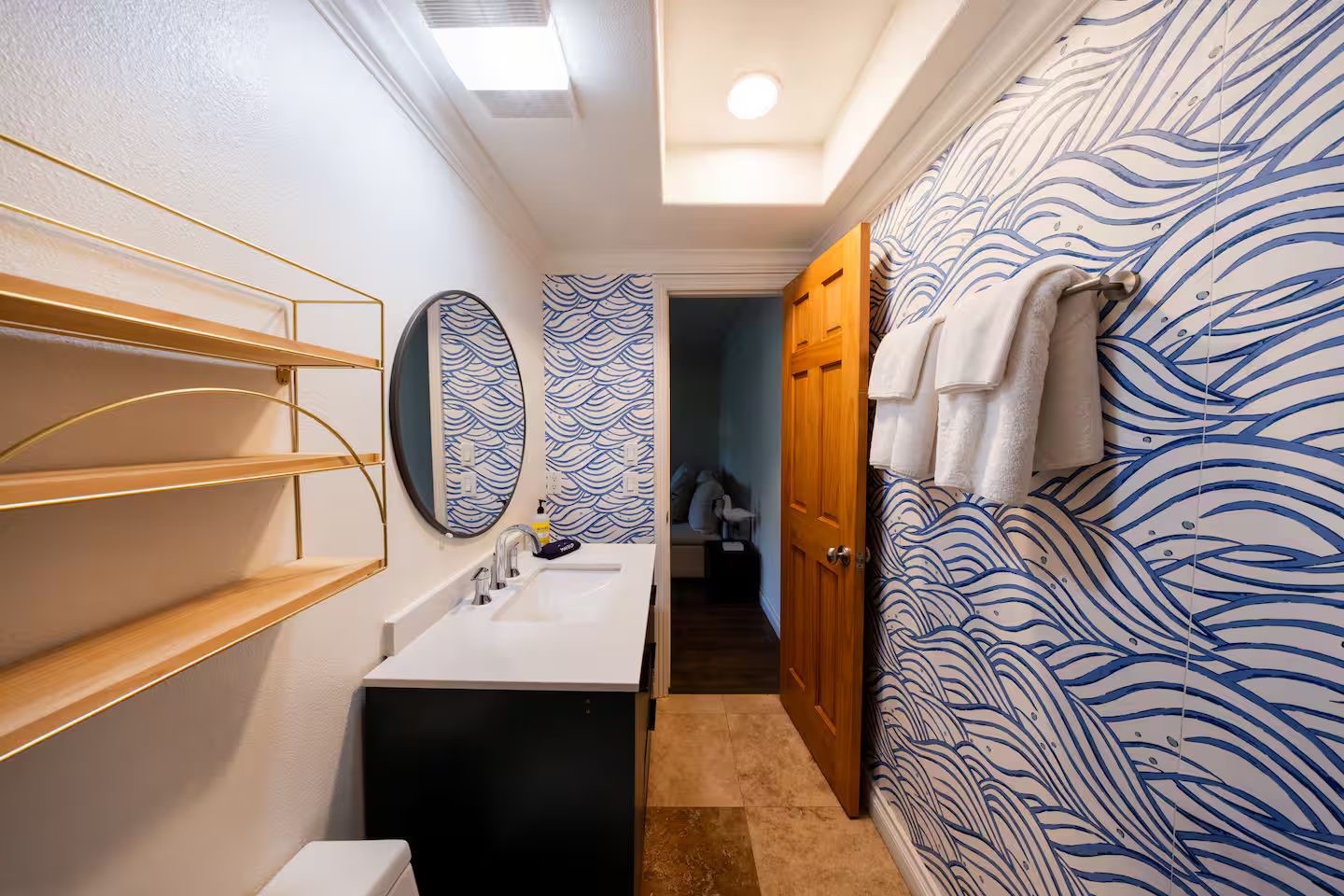 A bathroom with a white sink, mirror, and shelves, a toilet, and a shower area with a patterned wall.