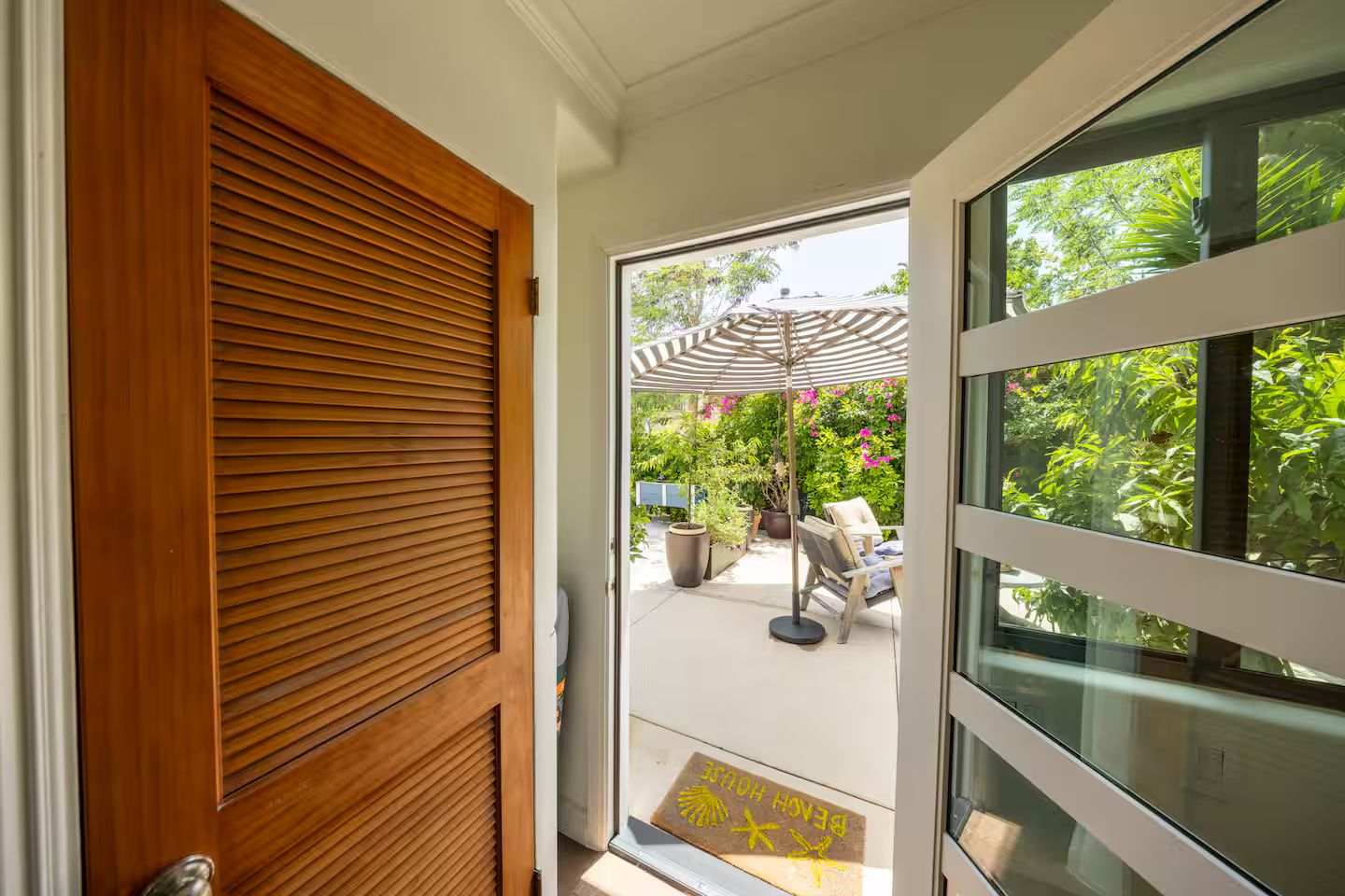A wooden door leading to an outdoor patio with a table and chairs, surrounded by lush greenery and a pergola.