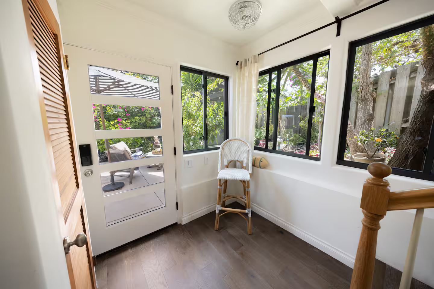 A room with a wooden floor, a white chair, a dining table with chairs, and a large window with a view of greenery.