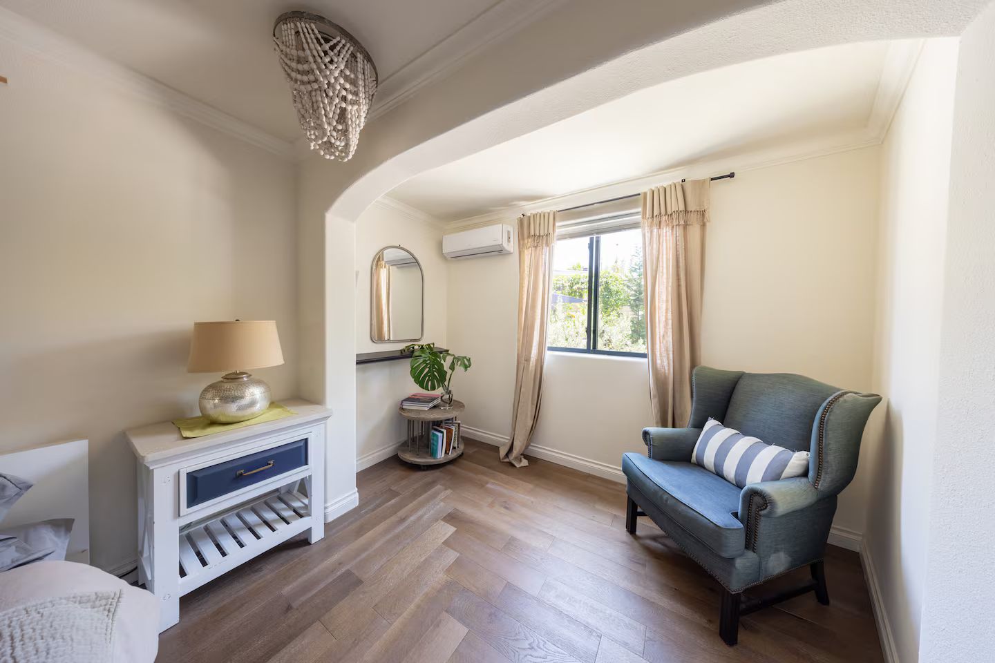 A room with a wooden floor, a blue armchair, a white console table with a lamp, a mirror, and a window with curtains.
