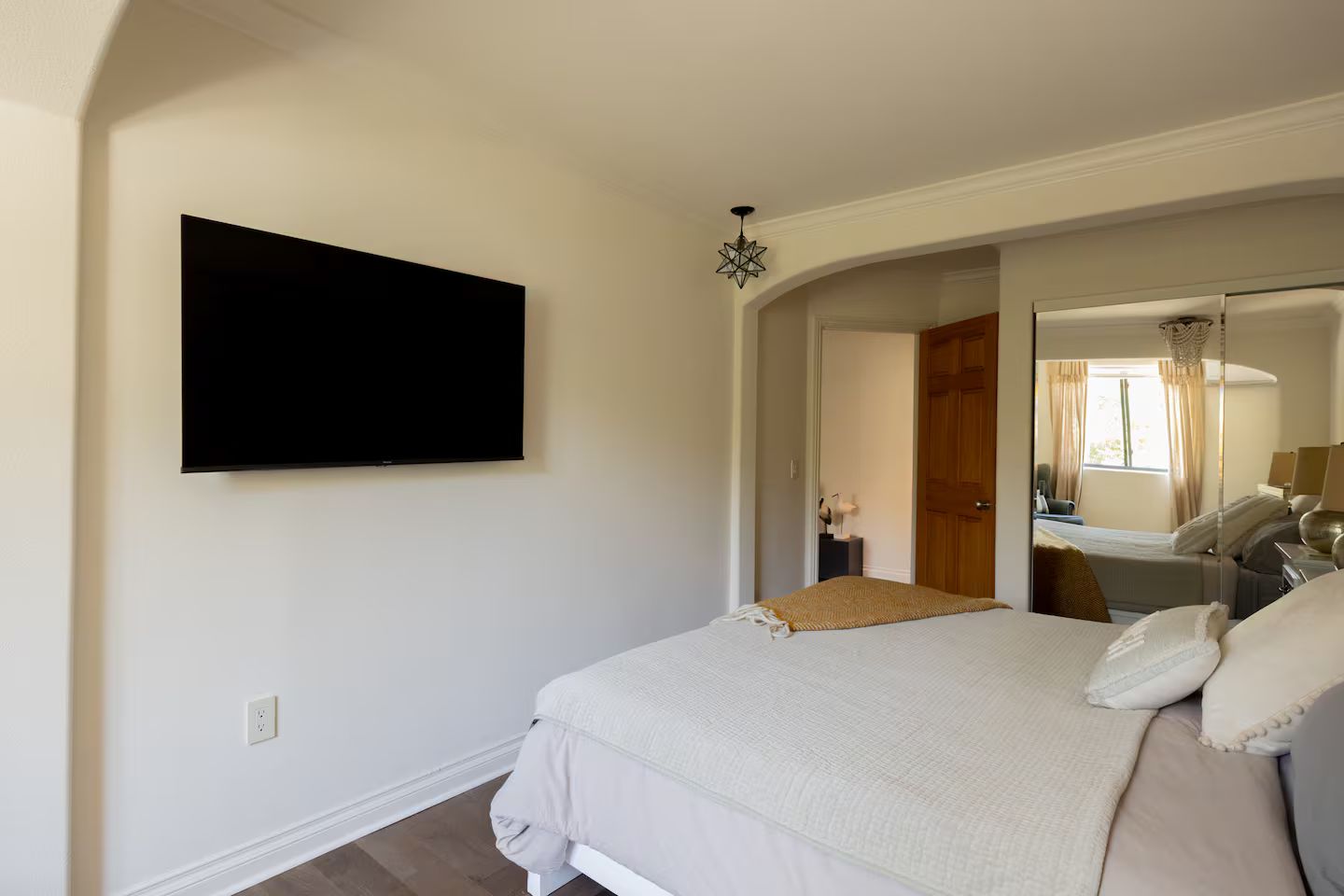 A bedroom with a bed, a flat-screen TV mounted on the wall, a bedside table with a lamp, and a mirror reflecting the room.