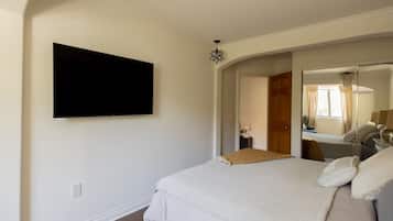 A bedroom with a bed, a flat-screen TV mounted on the wall, a bedside table with a lamp, and a mirror reflecting the room.