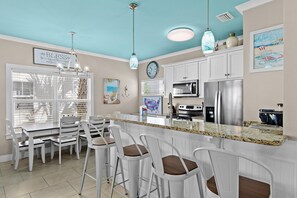 A kitchen with white cabinetry, a granite countertop, a stainless steel refrigerator, and a dining area with white chairs. - White Sands Escape (Miramar Beach)
