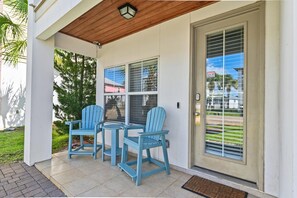 A covered porch with two chairs and a table, a door with a security camera warning, and a view of a residential area. - White Sands Escape (Miramar Beach)