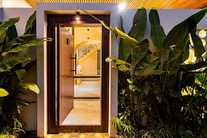 Uma entrada moderna com porta de madeira, escadaria interna e plantas tropicais em ambos os lados.