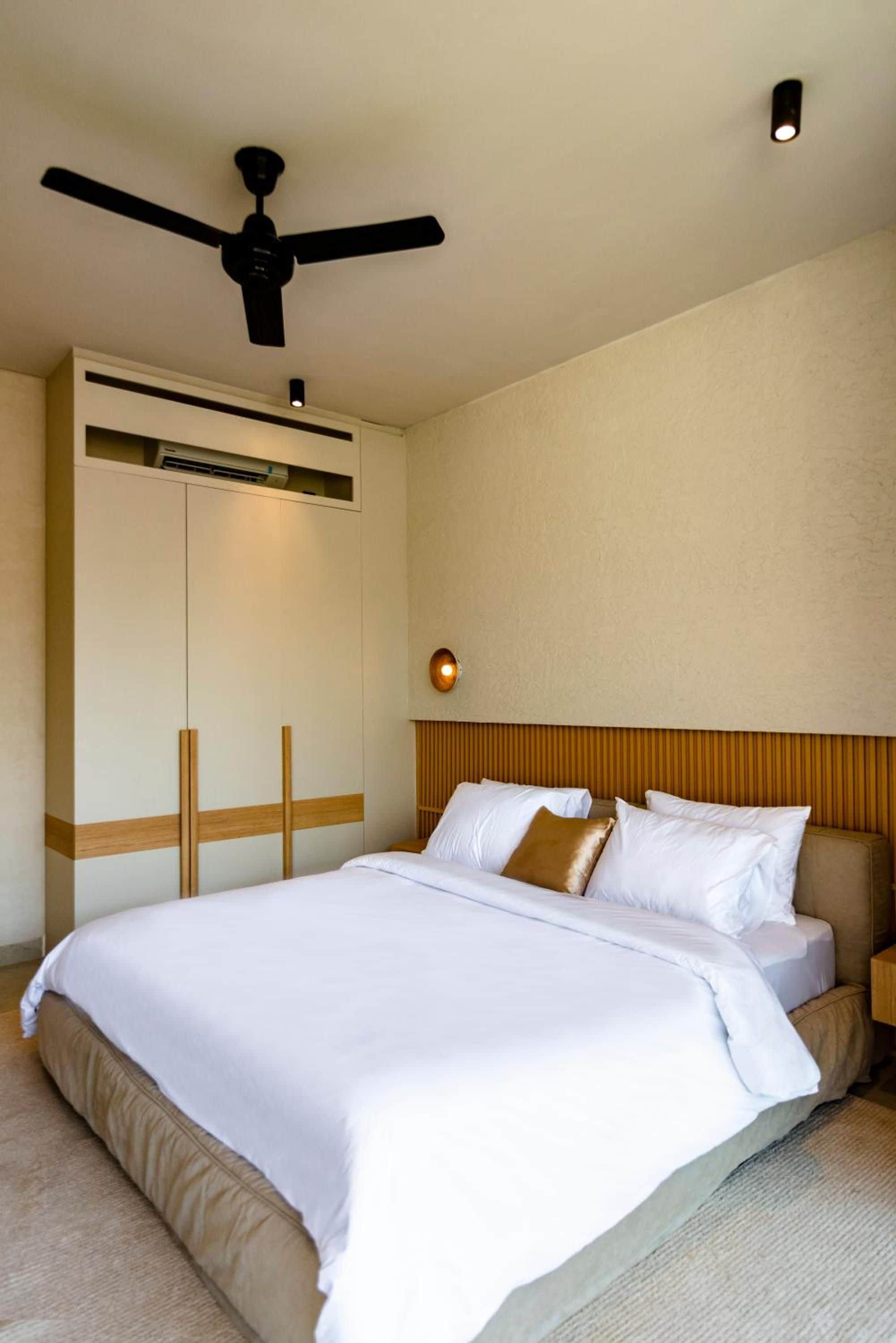 Una habitación de hotel con cama, ventilador de techo, aire acondicionado de pared y mesita de noche con lámpara.