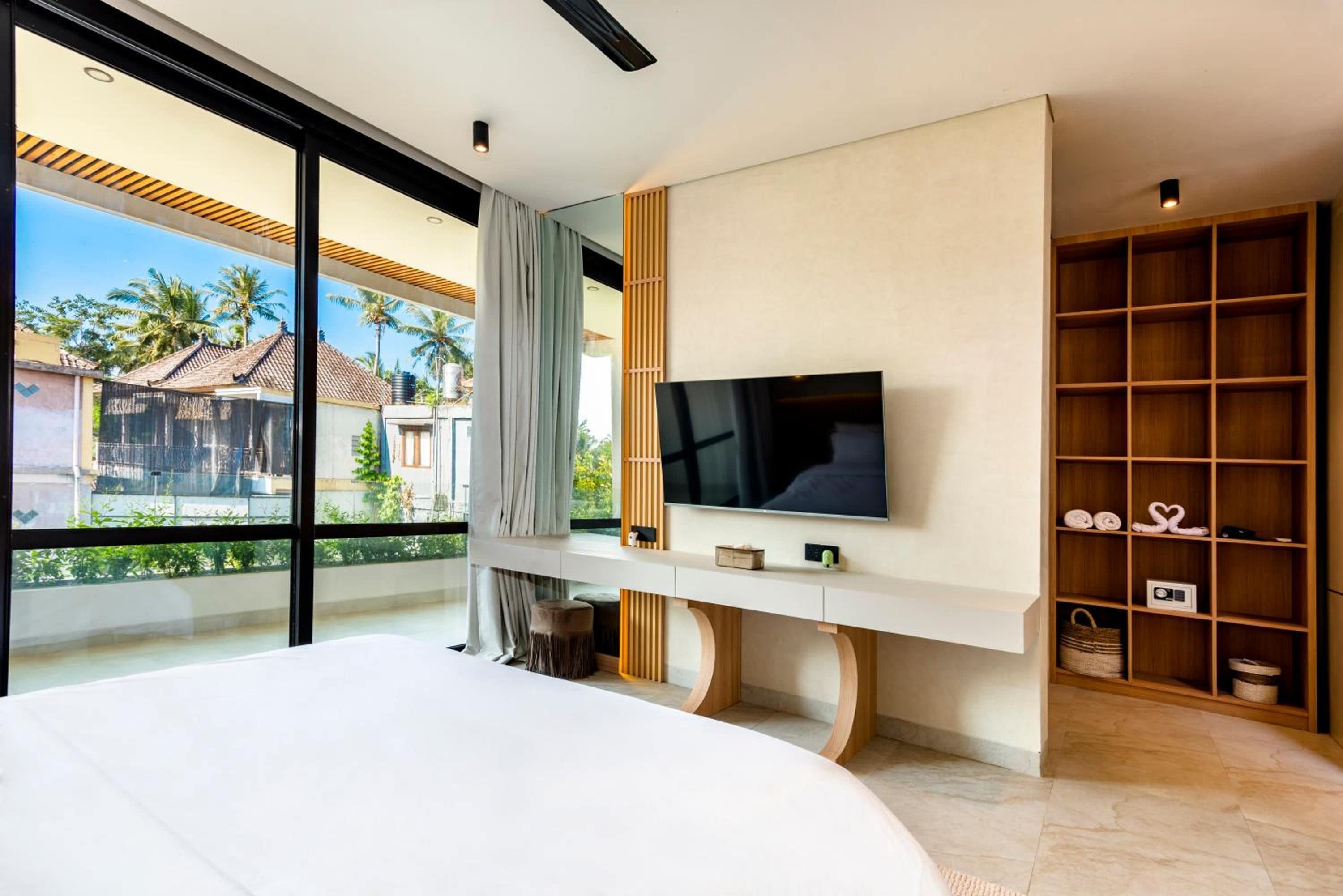 Un dormitorio moderno con una cama grande, un televisor montado en un mueble blanco y vistas a palmeras a través de amplias ventanas.