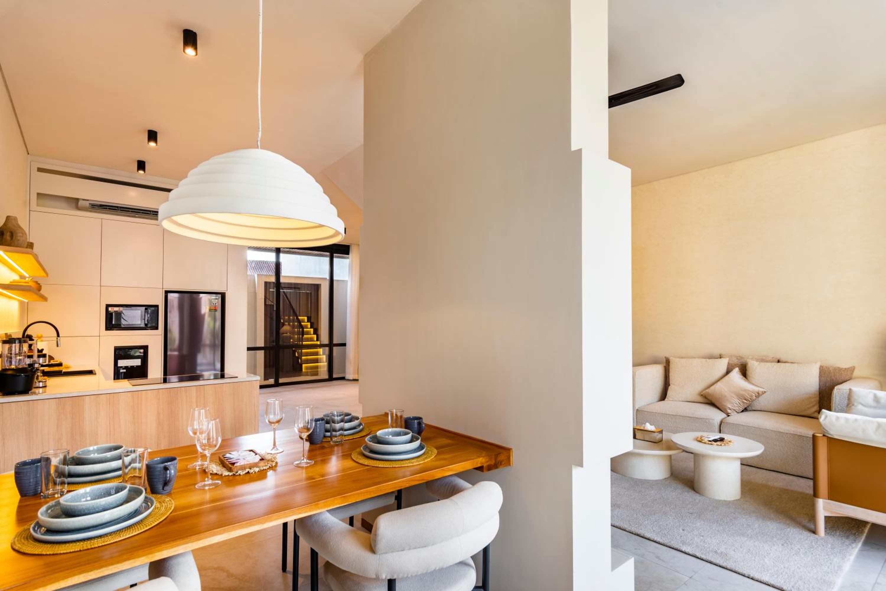 A modern kitchen with a wooden dining table, white pendant lighting, and a view of a staircase leading to a living area.