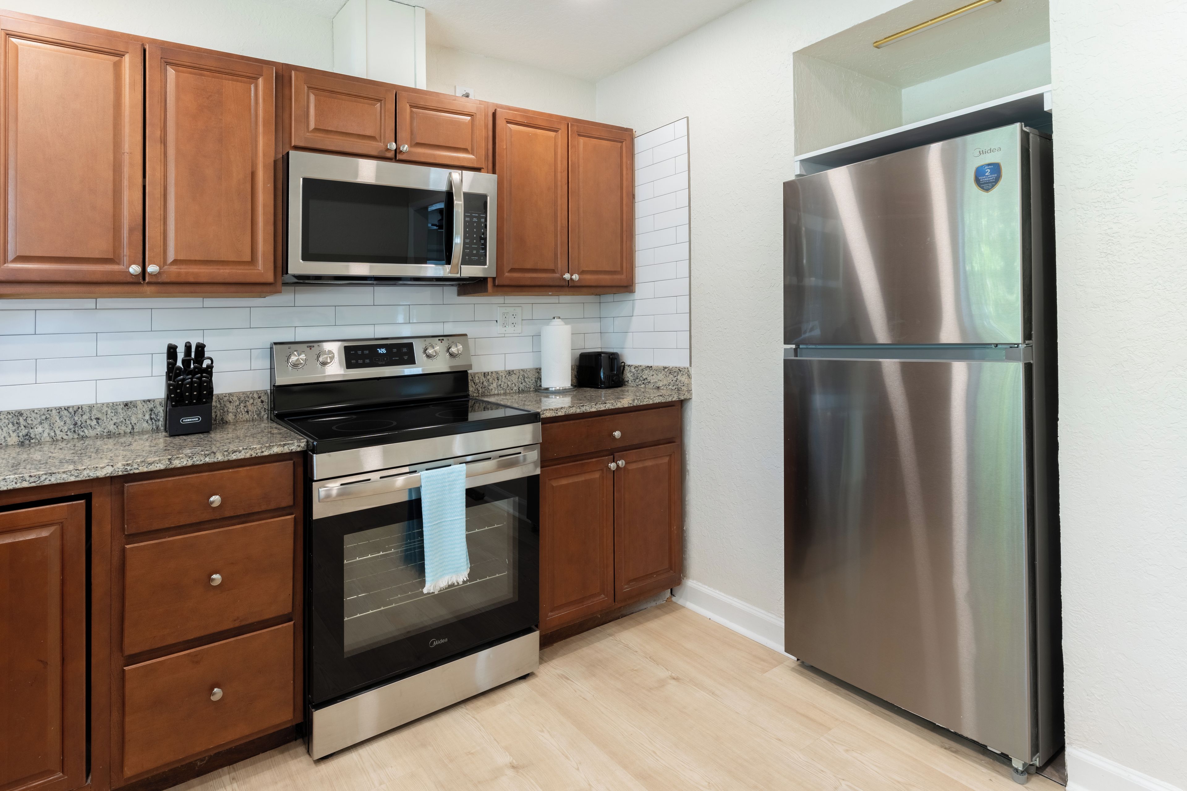 A kitchen with wooden cabinets, a stainless steel refrigerator, a microwave, an oven, and a granite countertop.