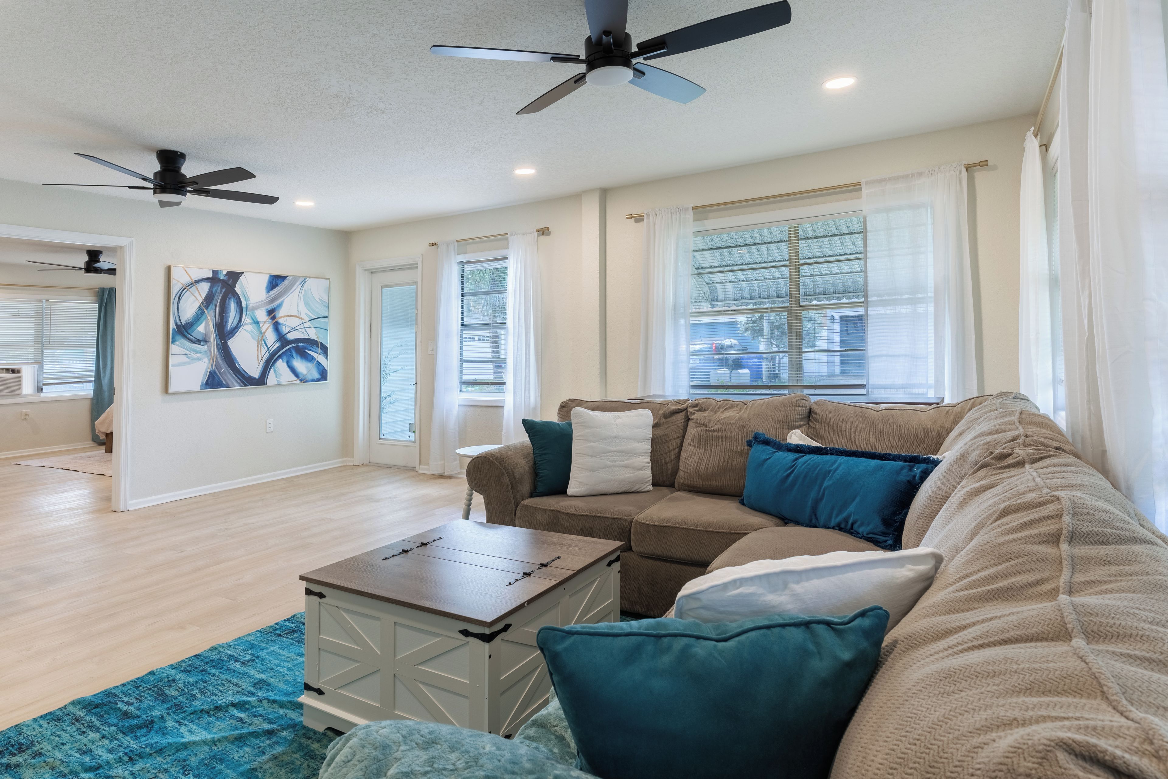 A spacious living room with a beige sofa, a coffee table, and a blue rug. There are ceiling fans, a large window with blinds, and a painting on the wall.