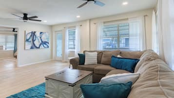 A spacious living room with a beige sofa, a coffee table, and a blue rug. There are ceiling fans, a large window with blinds, and a painting on the wall.