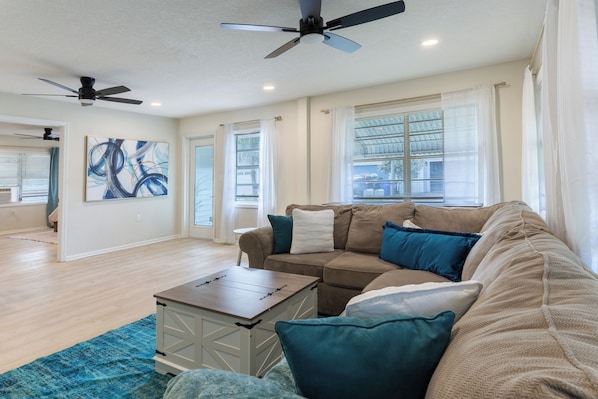 A spacious living room with a beige sofa, a coffee table, and a blue rug. There are ceiling fans, a large window with blinds, and a painting on the wall.