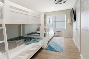 A room with bunk beds, a flat-screen TV, a window with blinds, a lamp, and a rug.