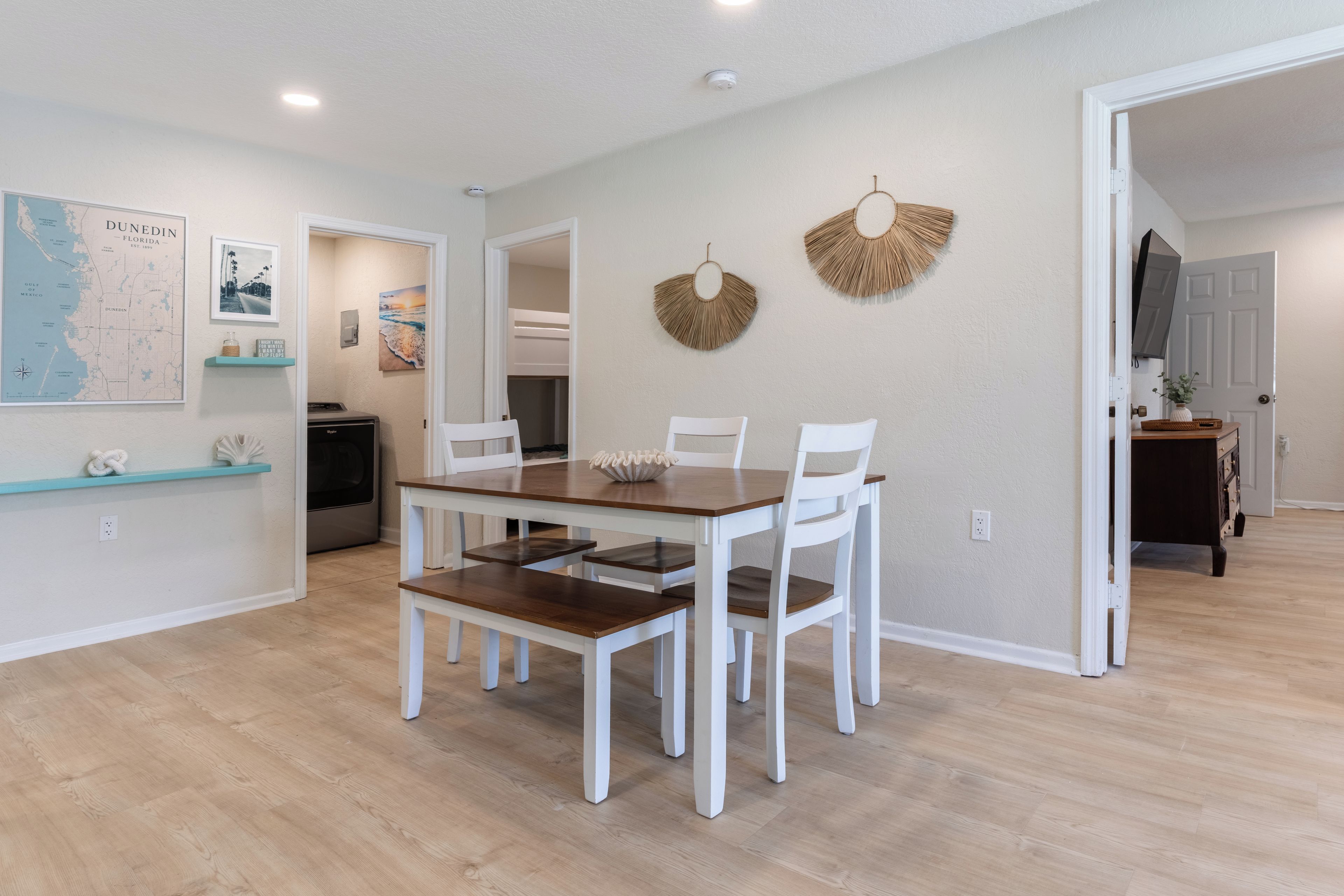 A dining area with a wooden table and white chairs, a map on the wall, and a kitchen in the background.