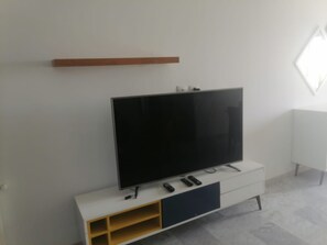 Uma sala de estar moderna com uma televisão de tela plana em um armário branco, uma prateleira de madeira acima e um radiador branco na parede.