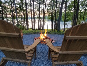 Zwei hölzerne Adirondack-Stühle, die ein Lagerfeuer mit einem See und Bäumen im Hintergrund zeigen.