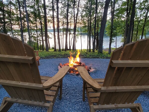 Two wooden Adirondack chairs facing a campfire with a lake and trees in the background.
