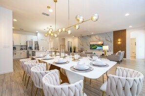 A modern dining area with a white table set for six, surrounded by plush chairs, a kitchen in the background, and a large TV mounted on the wall. - The best 9-suite house at Sonoma Resort (Kissimmee)