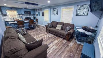 A living room with a sofa set, a coffee table, and a dining area with chairs. There is a kitchen in the background with a microwave and a coffee machine.