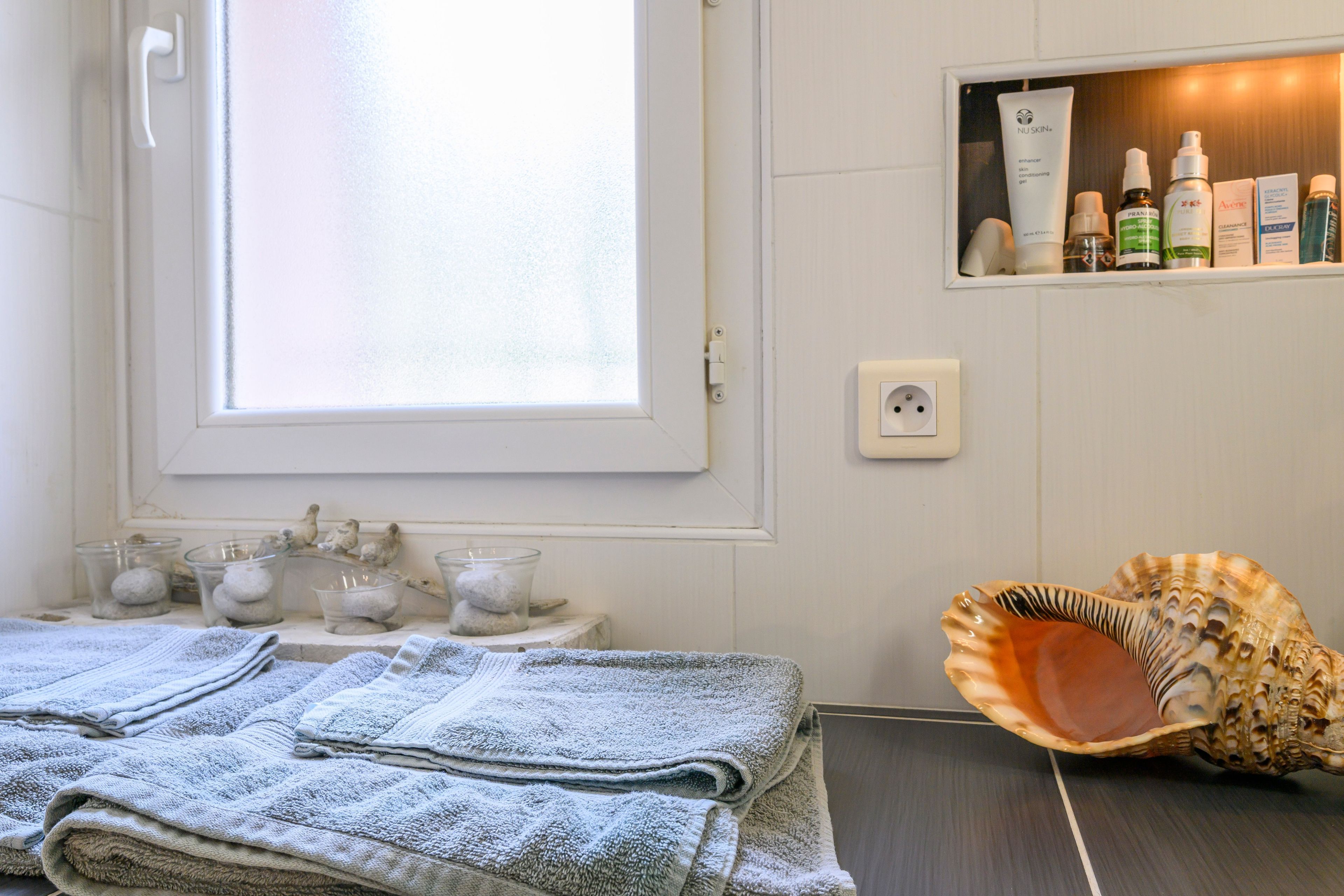 A bathroom with a window, a shelf with toiletries, and a decorative shell.