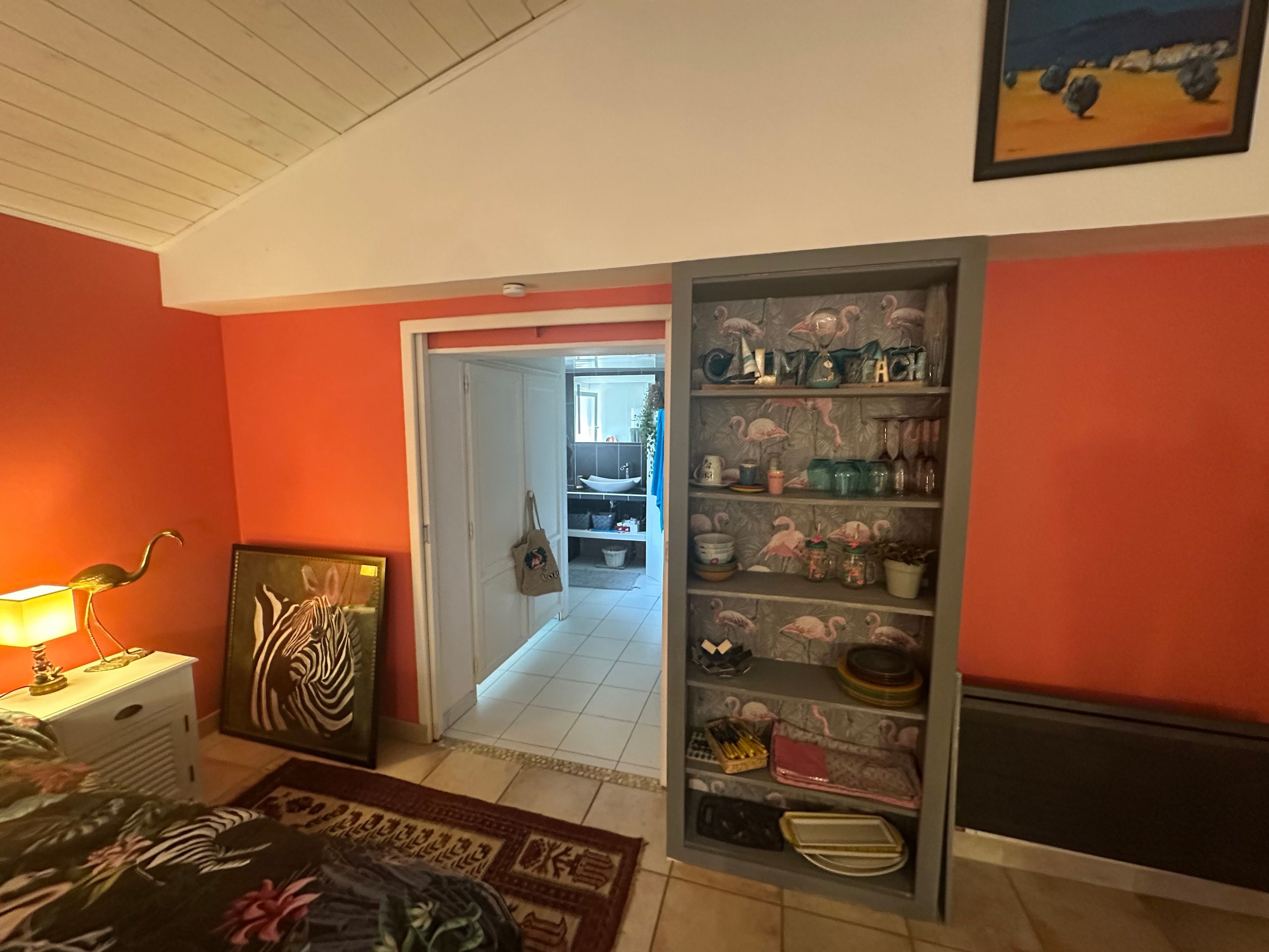 A room with a bed, a lamp, a painting, a shelf with decorative items, and a view into another room.