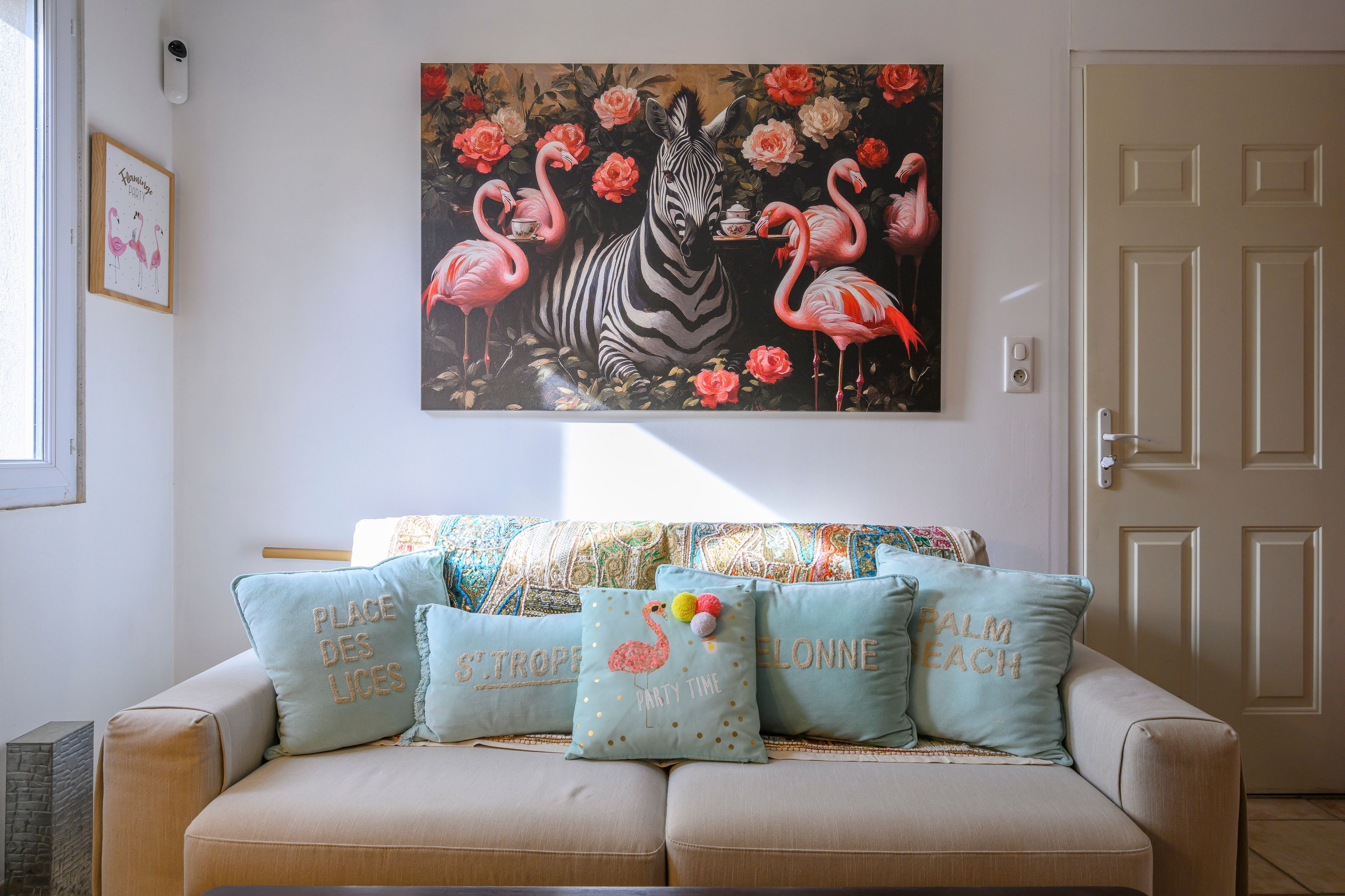 En stue med en sofa pyntet med dekorative puder, et billede af flamingoer og en zebra på væggen, samt en dør med lyskontakt.