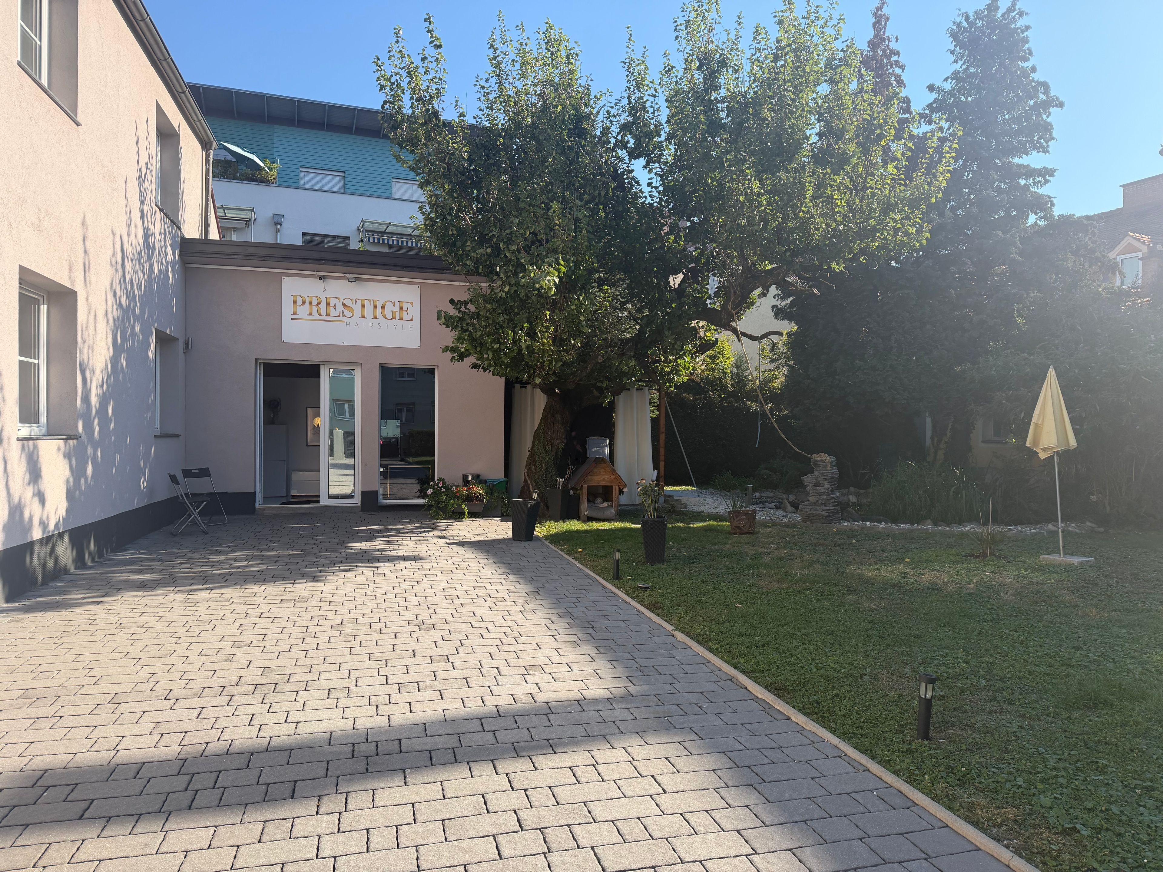 A building with a sign reading 'PRESTIGE HAIRSTYLE' and a paved walkway leading to a grassy area with a tree and a small structure.