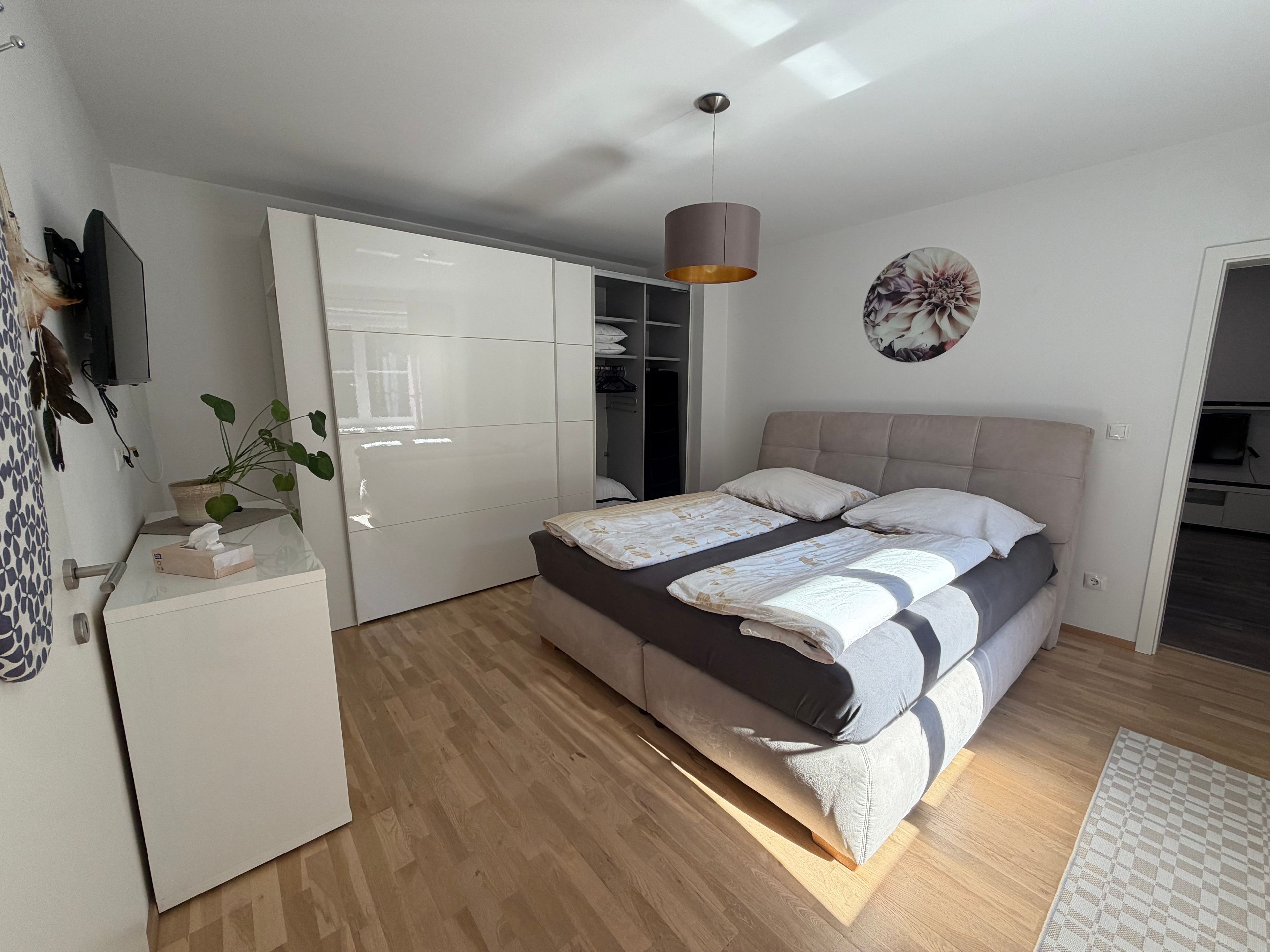 A bedroom with a bed, bedside tables, a wardrobe, a plant, and a wall-mounted TV.