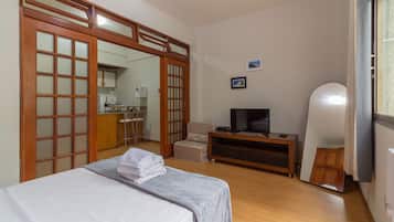 A hotel room with a bed, a television, a small desk, a chair, and a kitchen area visible through an open door.