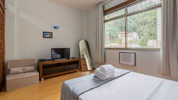 A hotel room with a bed, a TV on a wooden stand, a chair, and a window with a view of greenery.