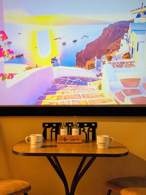 A television displaying a scenic view of a coastal village with boats, a wooden table with coffee mugs, and a coffee syrup dispenser. - Theatre Suite in Deep Ellum - Cinema, Skyline & City Vibes (Dallas)