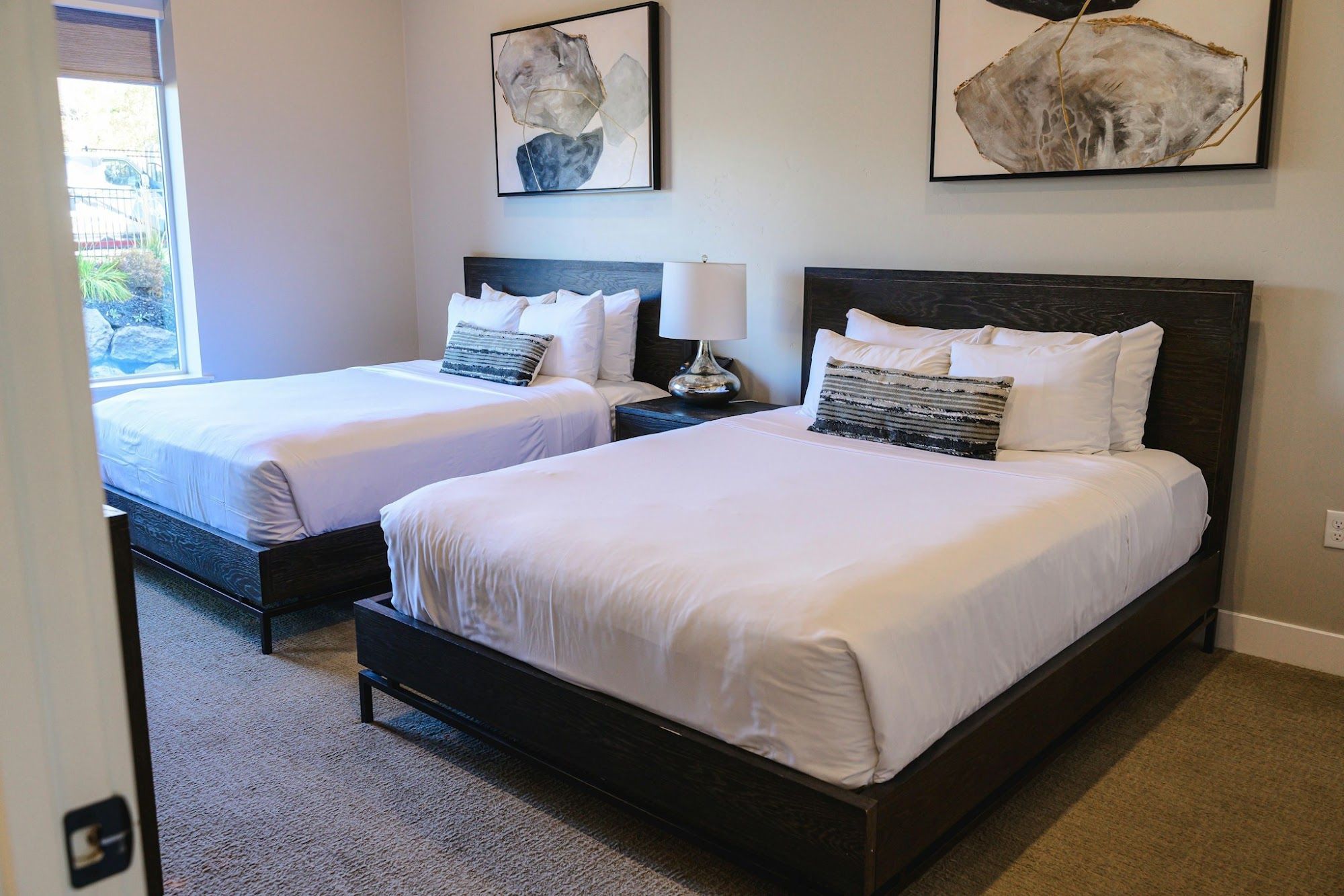A hotel room with two beds, a nightstand with a lamp, and artwork on the walls.
