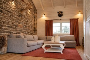 A cozy living room with a stone wall, a sectional sofa, a white coffee table, and a window with curtains. - VALENTINA LODGE – Luxury vacation home near Durbuy with a spa area and game room (Durbuy)