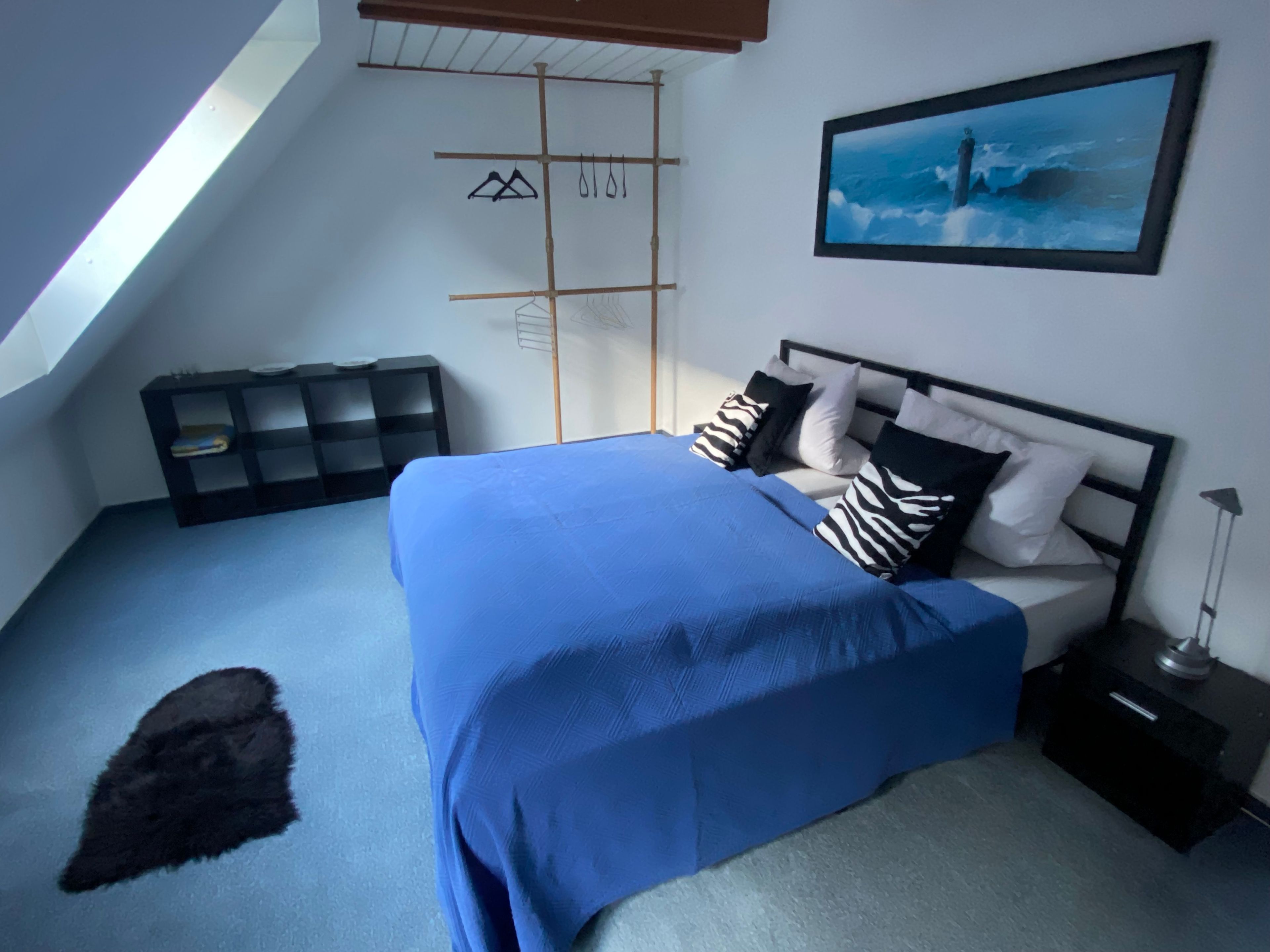 Une chambre avec un plafond en pente, un lit recouvert d’une couverture bleue, un oreiller à motifs noir et blanc, une table de chevet noire et un tableau encadré accroché au mur.