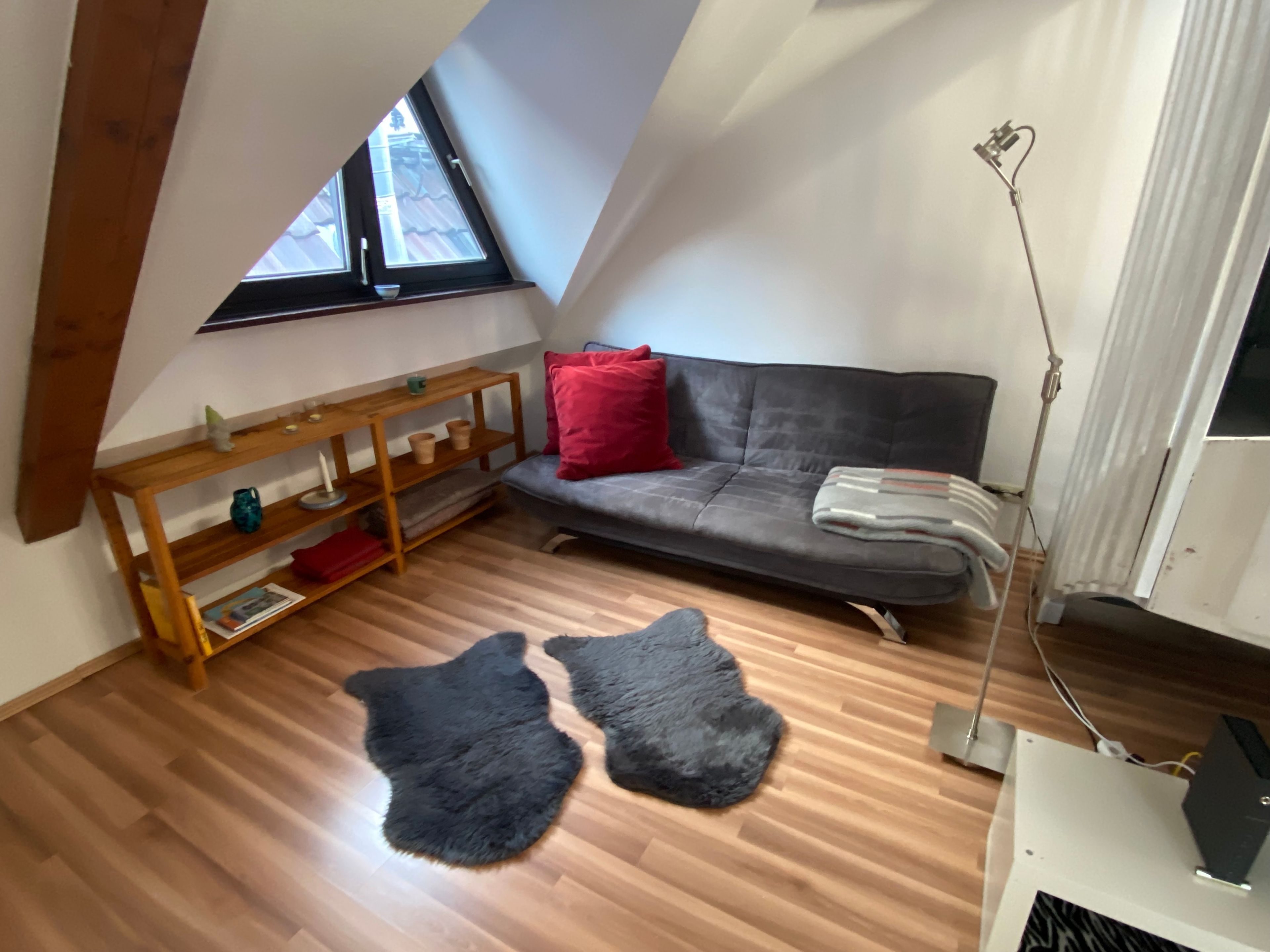 Ett rum i loftstil med ett sluttande tak, en grå soffa, ett bord i trä, en svart, lurvig matta och en golvlampa.