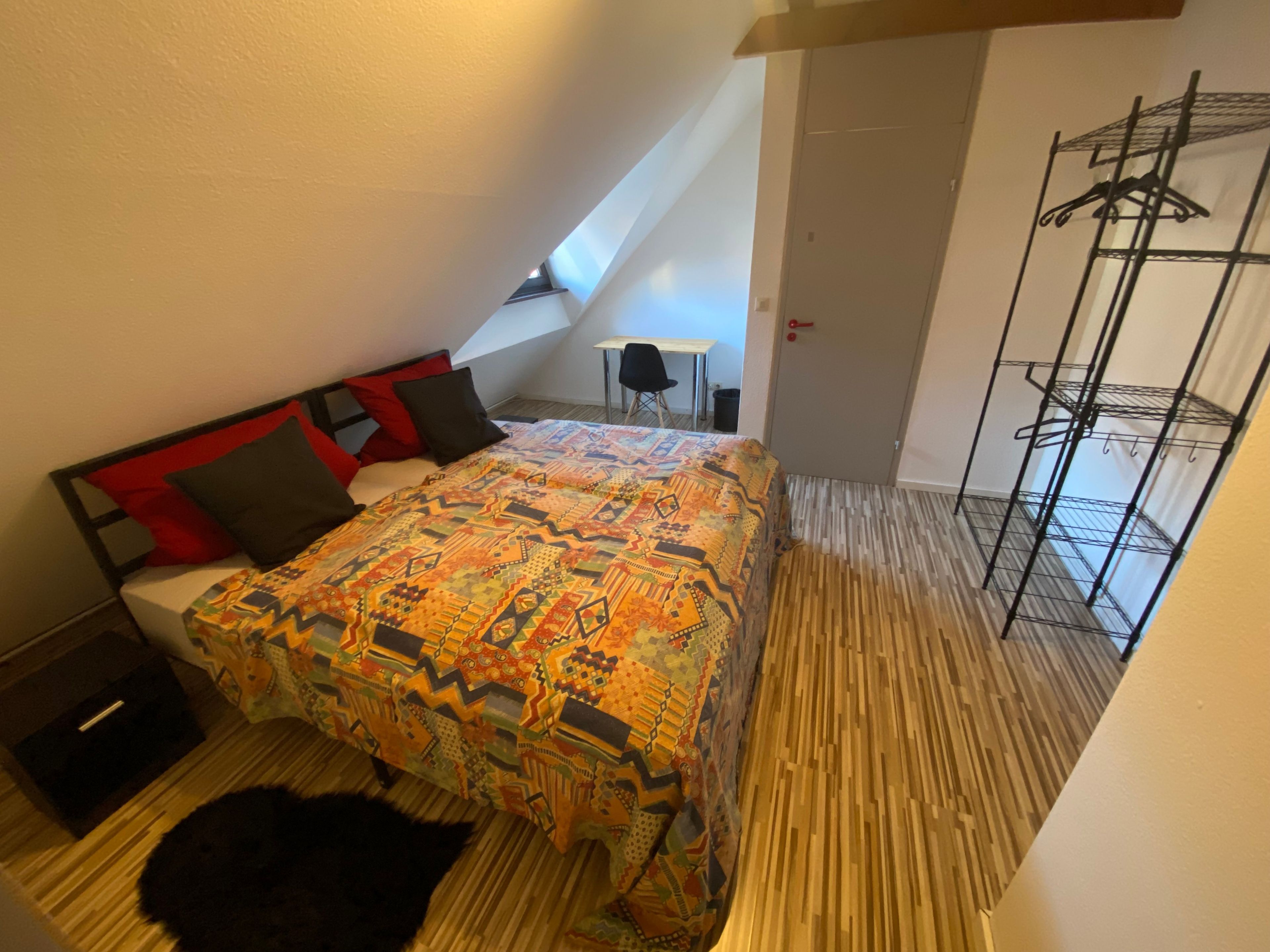 A bedroom with a bed, a black dog, a wooden floor, a window, and a clothing rack.