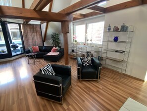 A spacious living area with wooden flooring, a black leather sofa, a black leather armchair, and a metal shelving unit against the wall.